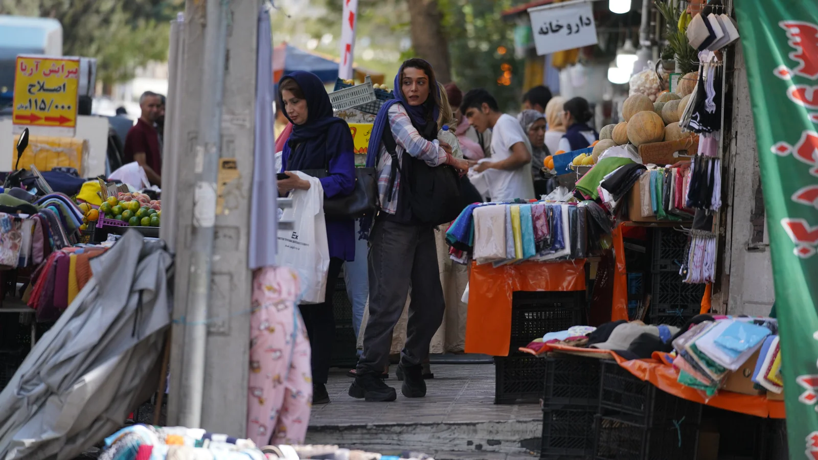 Calle de mercado con mujeres con pañuelo en la cabeza, puestos de fruta y telas, y un cartel en alfabeto árabe.