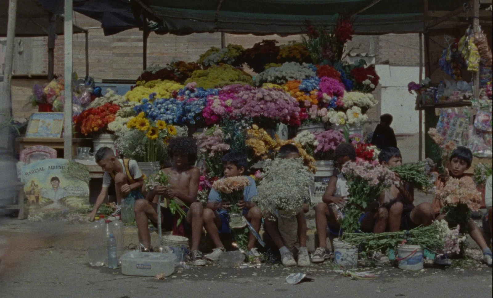 Varios niños sentados entre cubos y ramos de flores de colores, bajo una lona en una calle polvorienta.