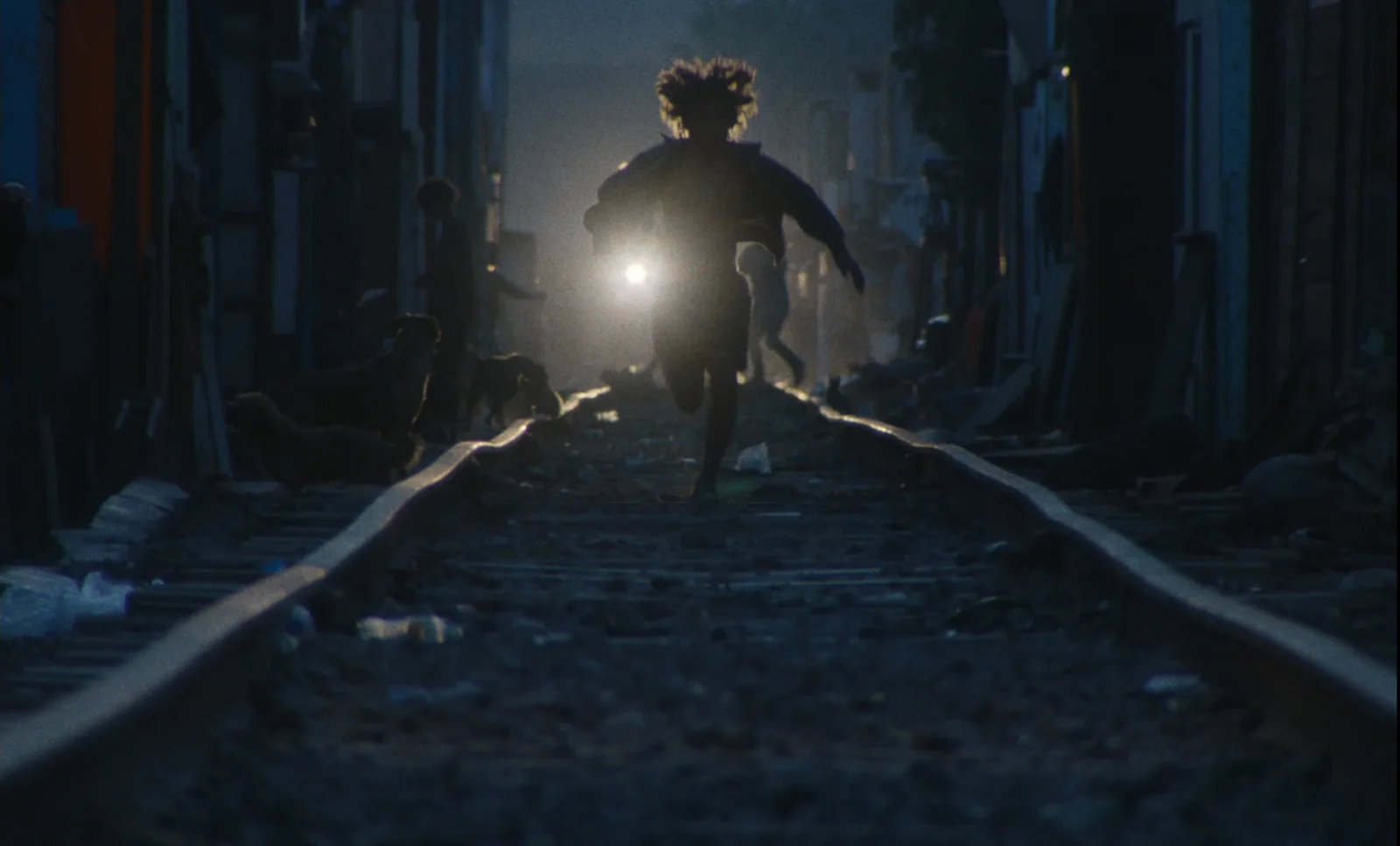 Silueta de un niño corriendo por unas vías de tren hacia una luz intensa, entre casas precarias.