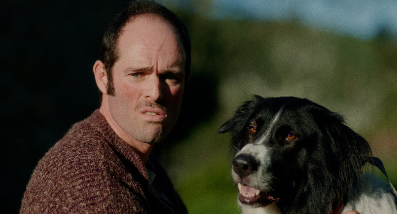 Hombre con jersey granate y gesto de desconfianza junto a un perro border collie blanco y negro.