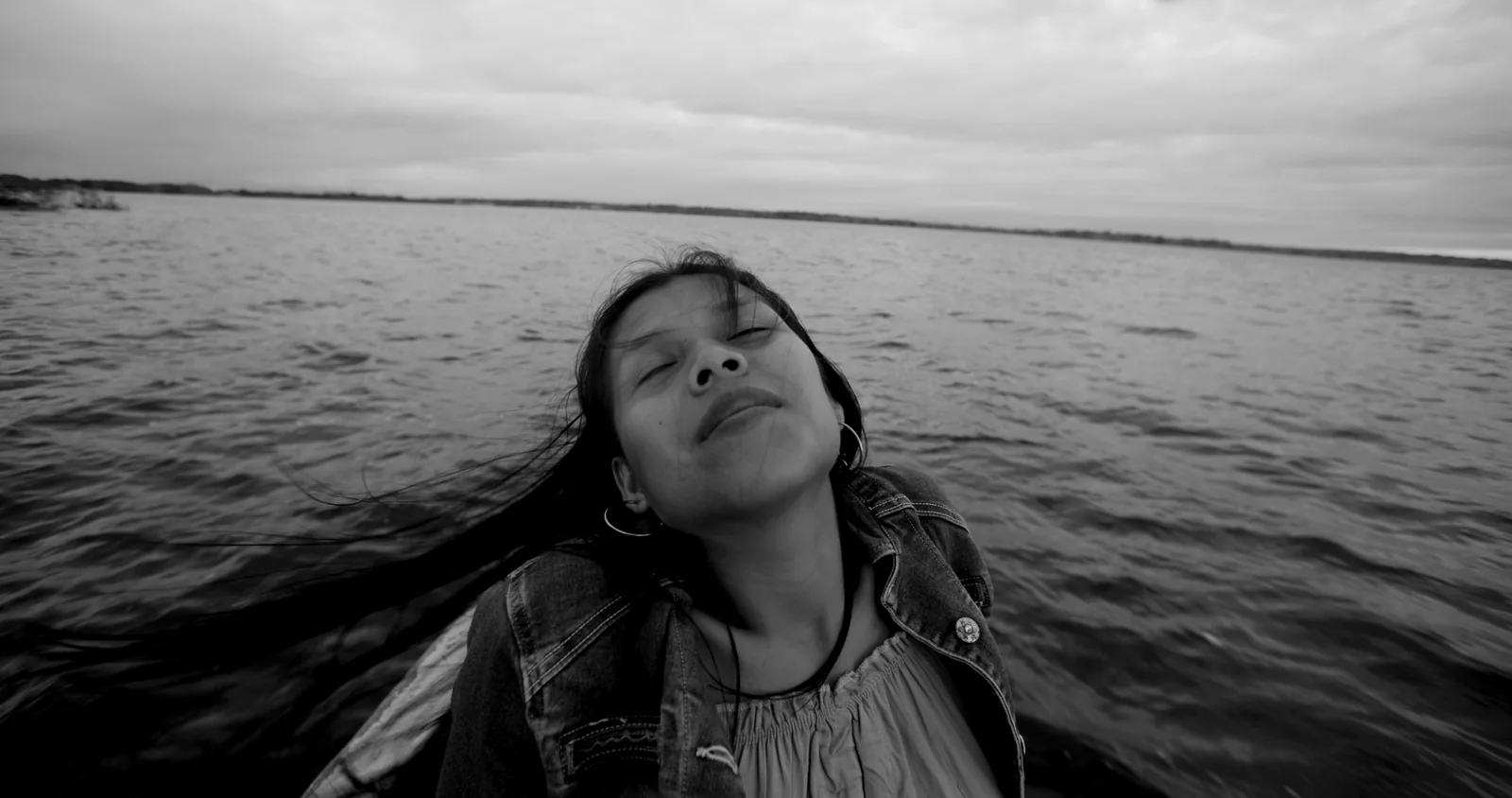 Blanco y negro de una joven con el pelo agitado por el viento y los ojos cerrados, sobre una superficie de agua.