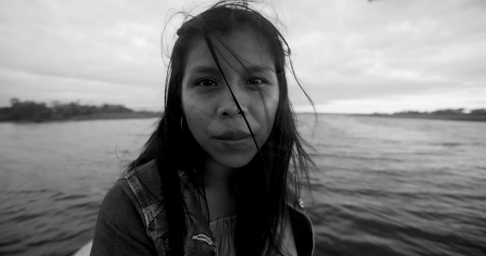 Blanco y negro de una joven de pelo largo y revuelto mirando a cámara, con el agua y el cielo detrás.