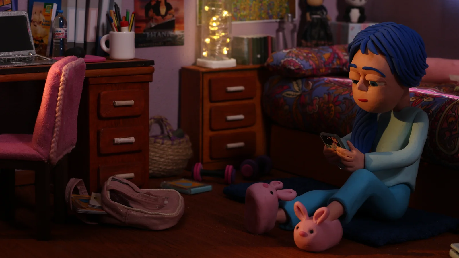 Animación en plastilina de una chica de pelo azul sentada en el suelo de su cuarto, mirando el móvil.