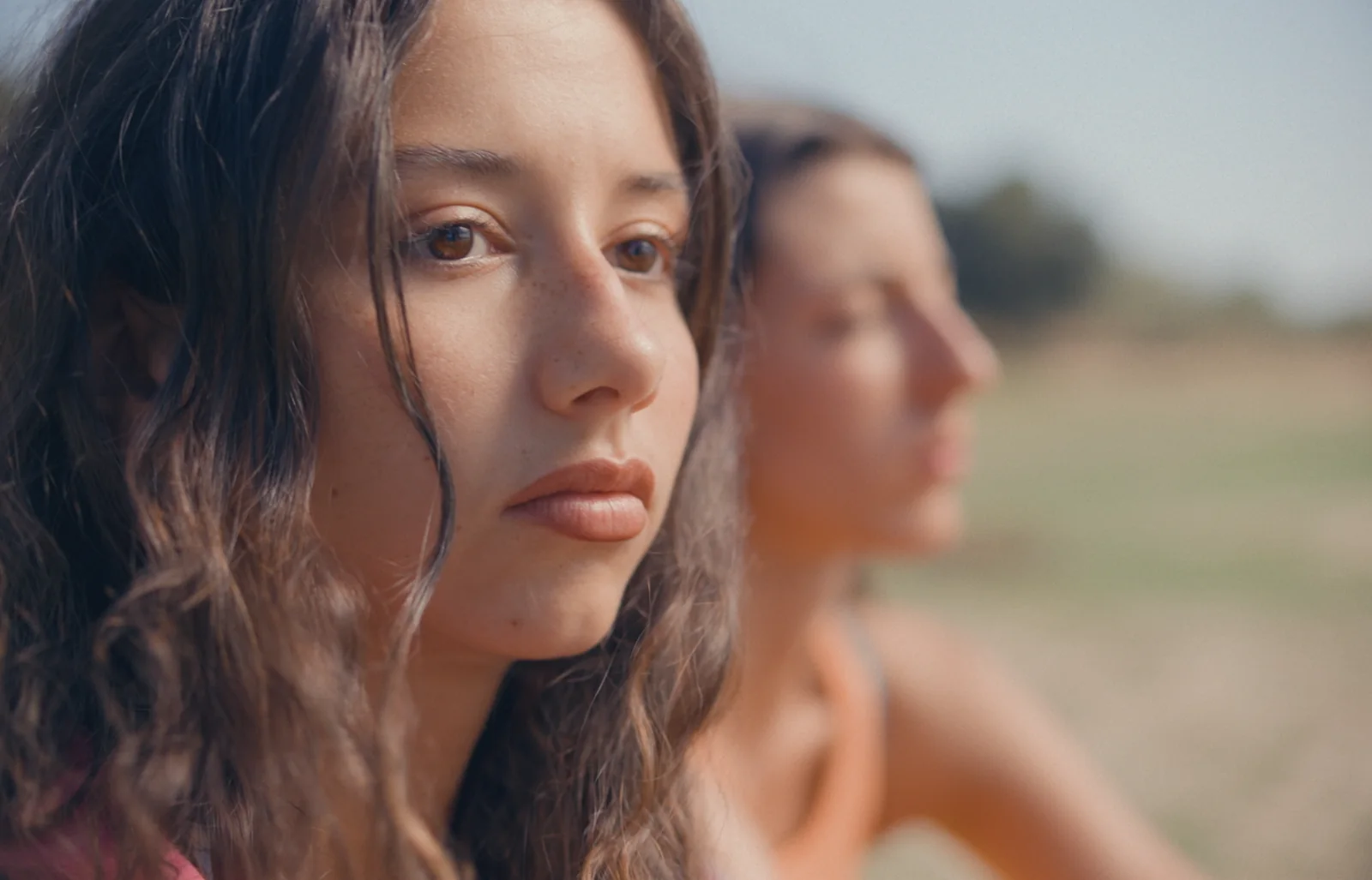 Primer plano de una joven de melena castaña y mirada seria, con otra mujer desenfocada detrás en un campo.