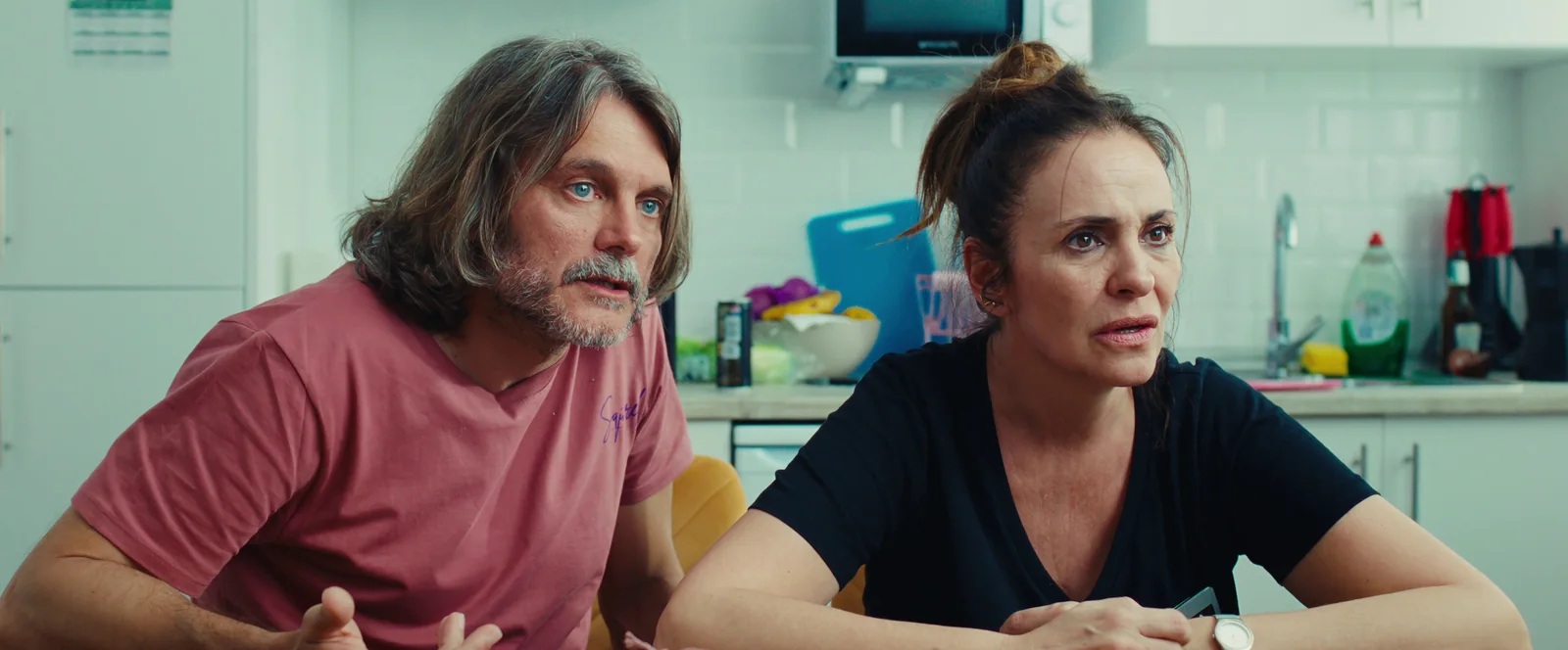 Pareja de mediana edad en una cocina blanca: él con camiseta rosa y barba, ella con moño y camiseta negra.