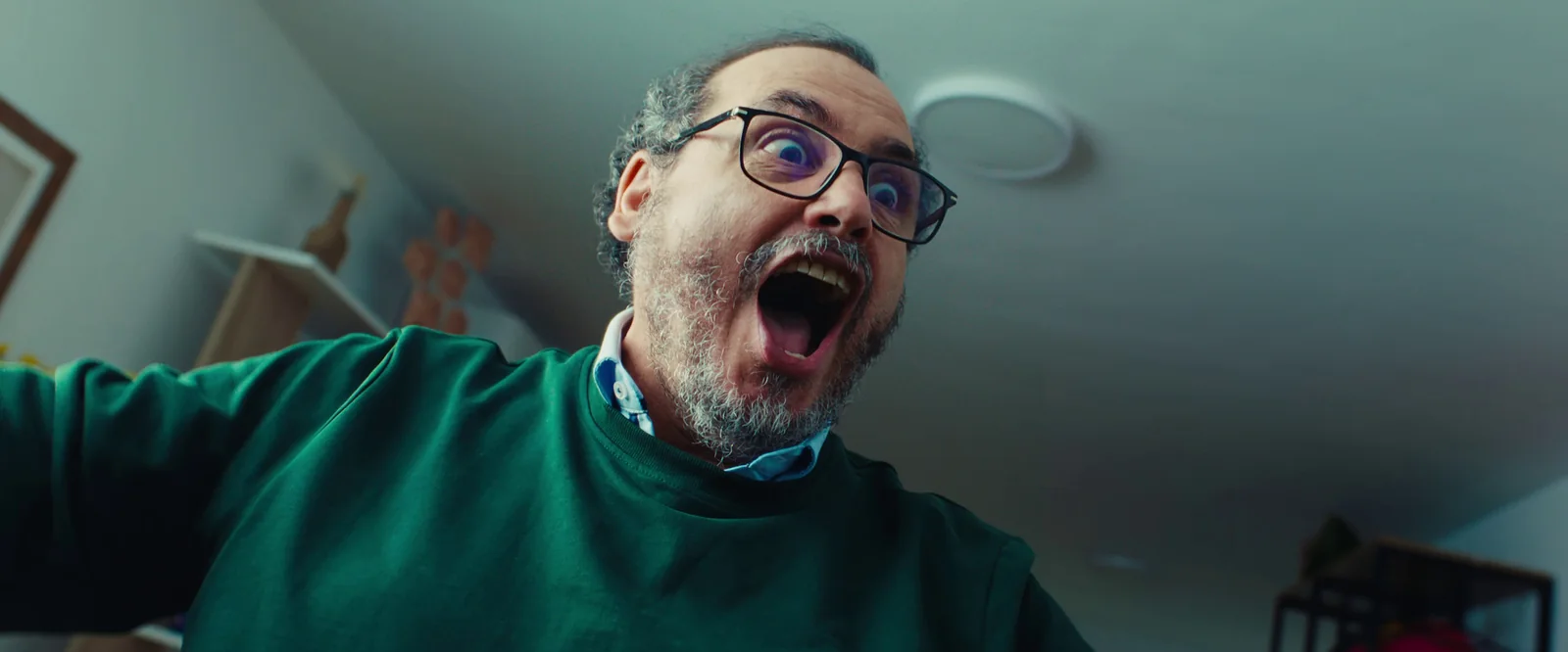 Hombre de mediana edad con gafas y jersey verde, con la boca abierta y gesto de sorpresa frente a la cámara.