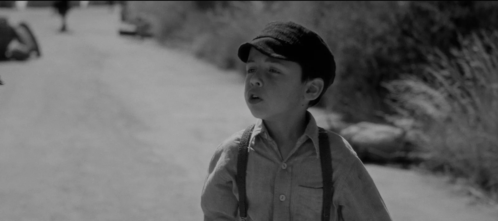 Niño pequeño en blanco y negro, con gorra y tirantes, caminando por un camino de tierra.