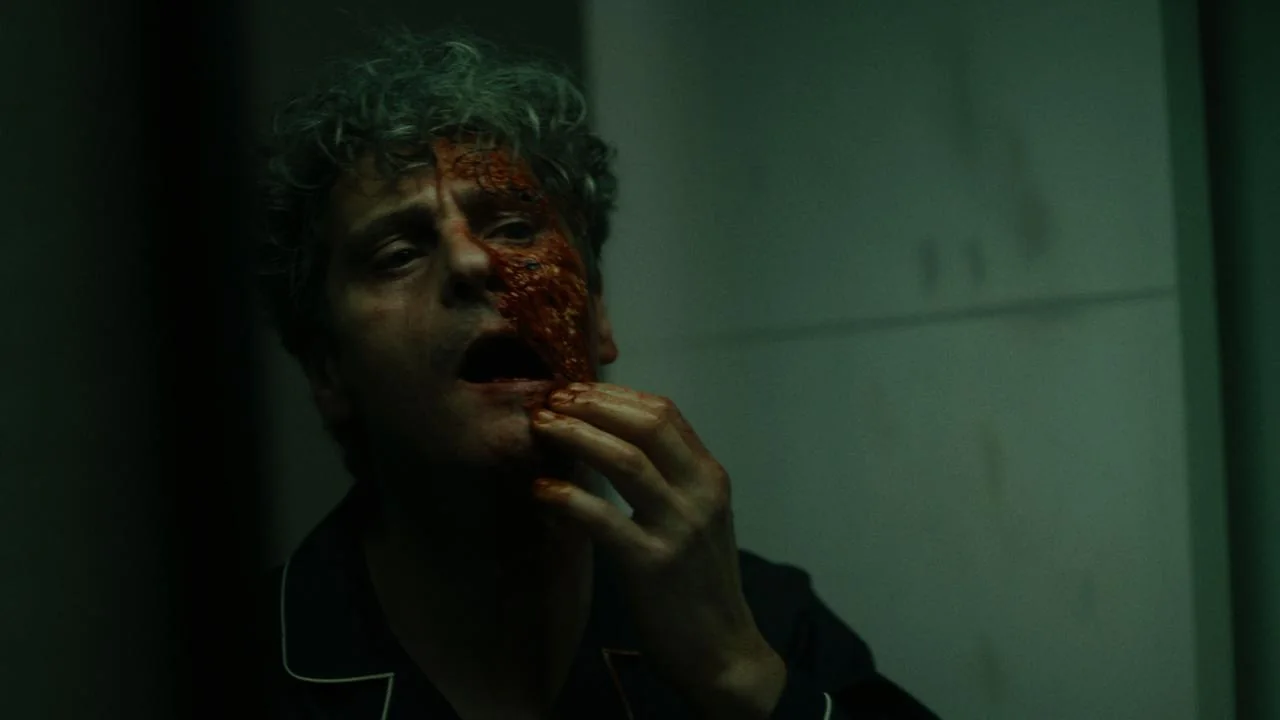 Hombre de pelo rizado con la mitad del rostro cubierta de sangre, tocándose la herida en un pasillo oscuro.
