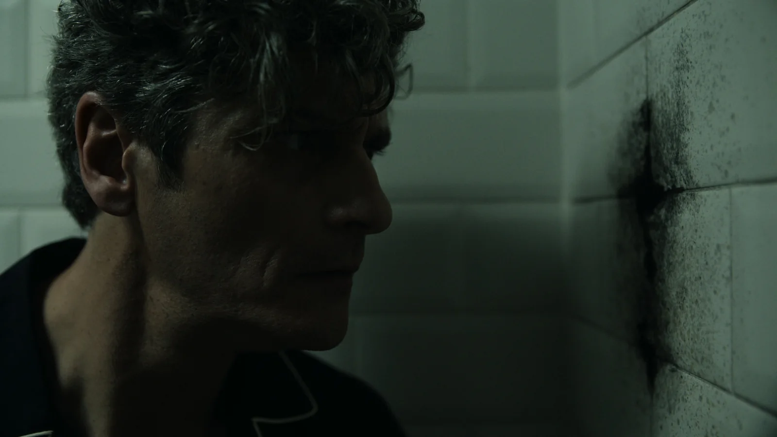 Hombre de perfil con pelo rizado, mirando una mancha oscura en una pared de azulejos blancos.