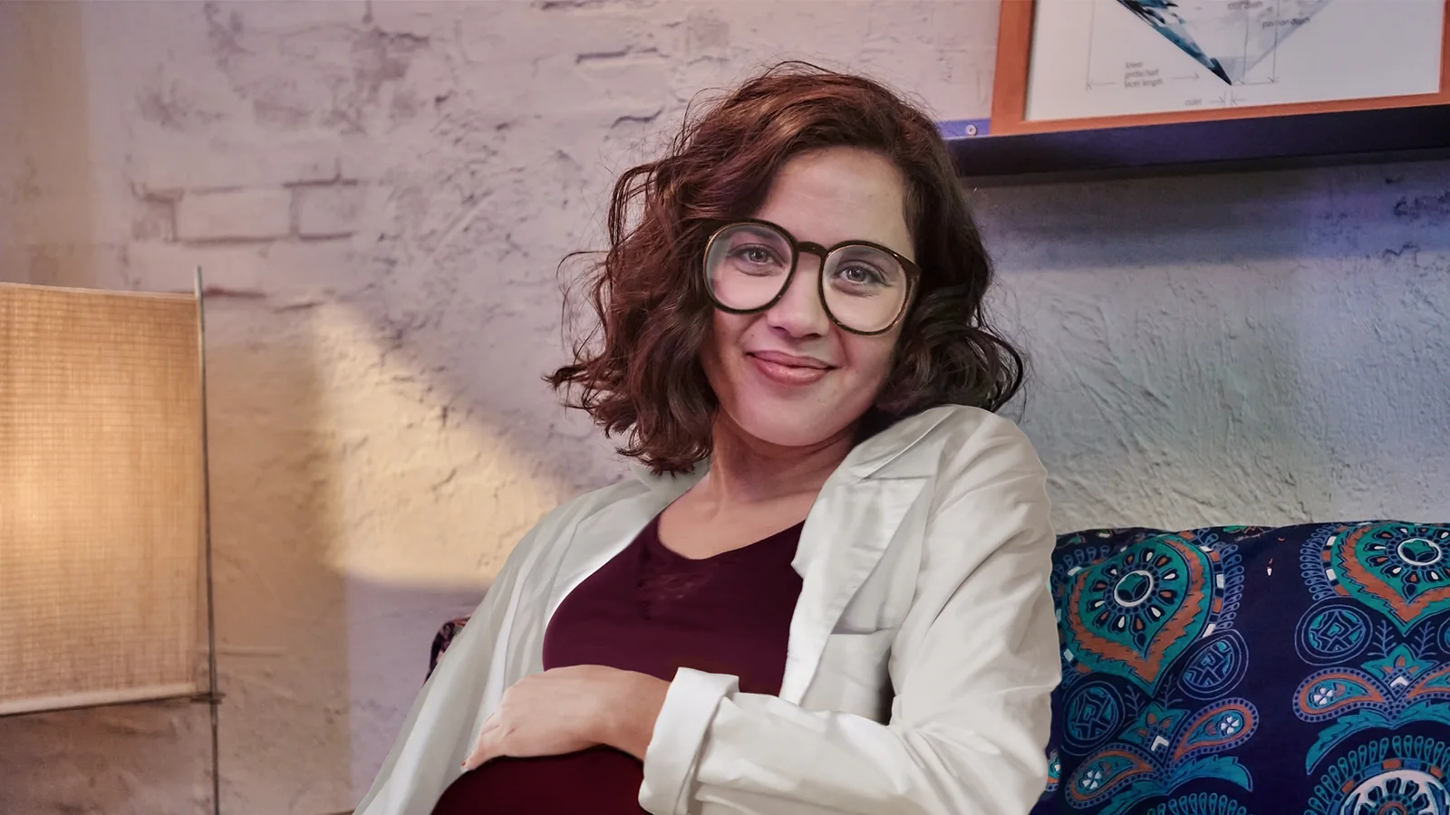 Mujer de pelo castaño rizado y gafas redondas, sonriendo, con la mano sobre el vientre y bata blanca.