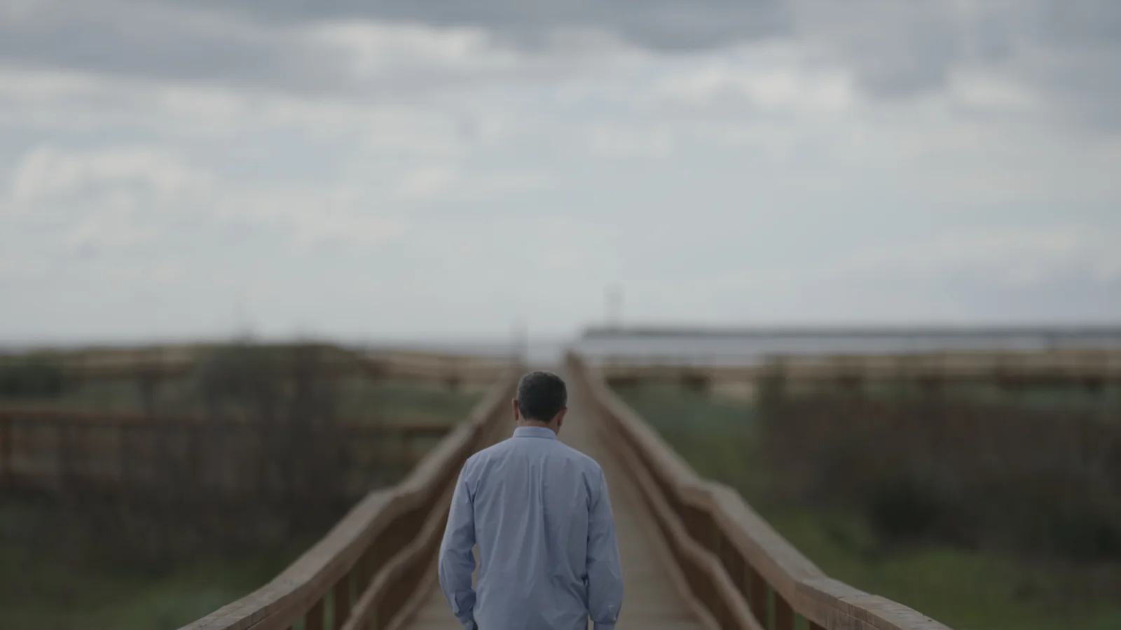 Hombre de espaldas, con camisa azul clara, caminando por una pasarela de madera hacia el horizonte.