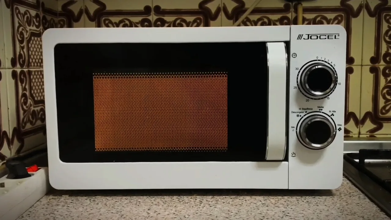Fotograma de «El microondas»: un microondas blanco sobre una encimera, encendido.