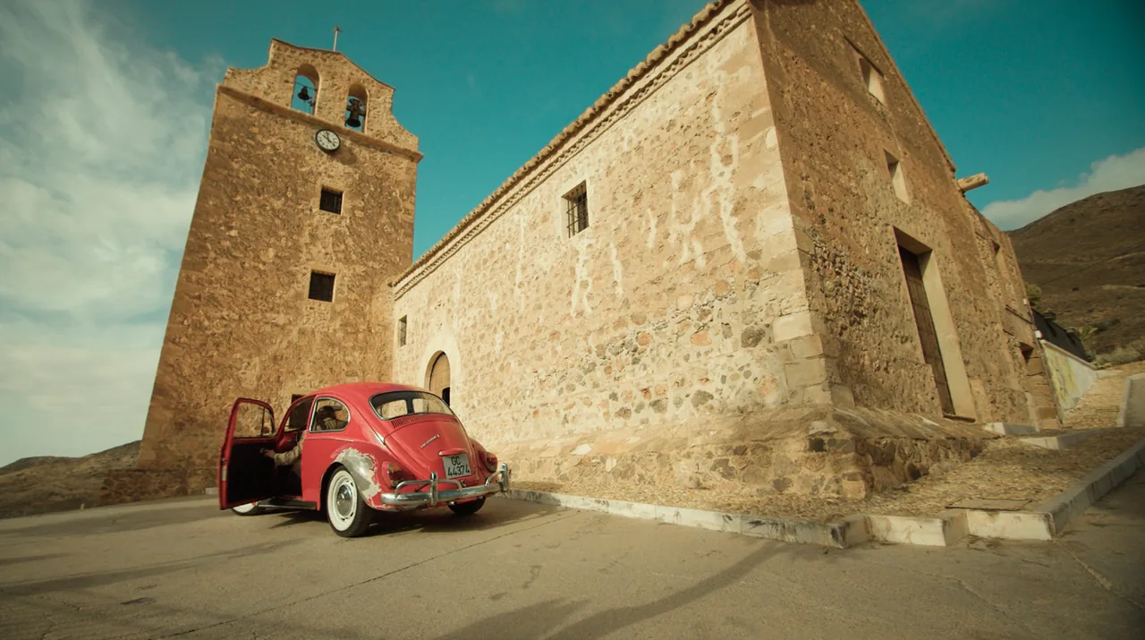 Fotograma de «Mariano Salvador España»: un coche rojo antiguo aparcado junto a una iglesia de piedra con torre.