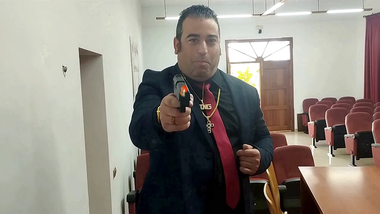 Fotograma de «Perdiendo la cabeza por La Viñuela»: un hombre con traje y corbata roja apunta con una pistola en un salón de actos vacío.