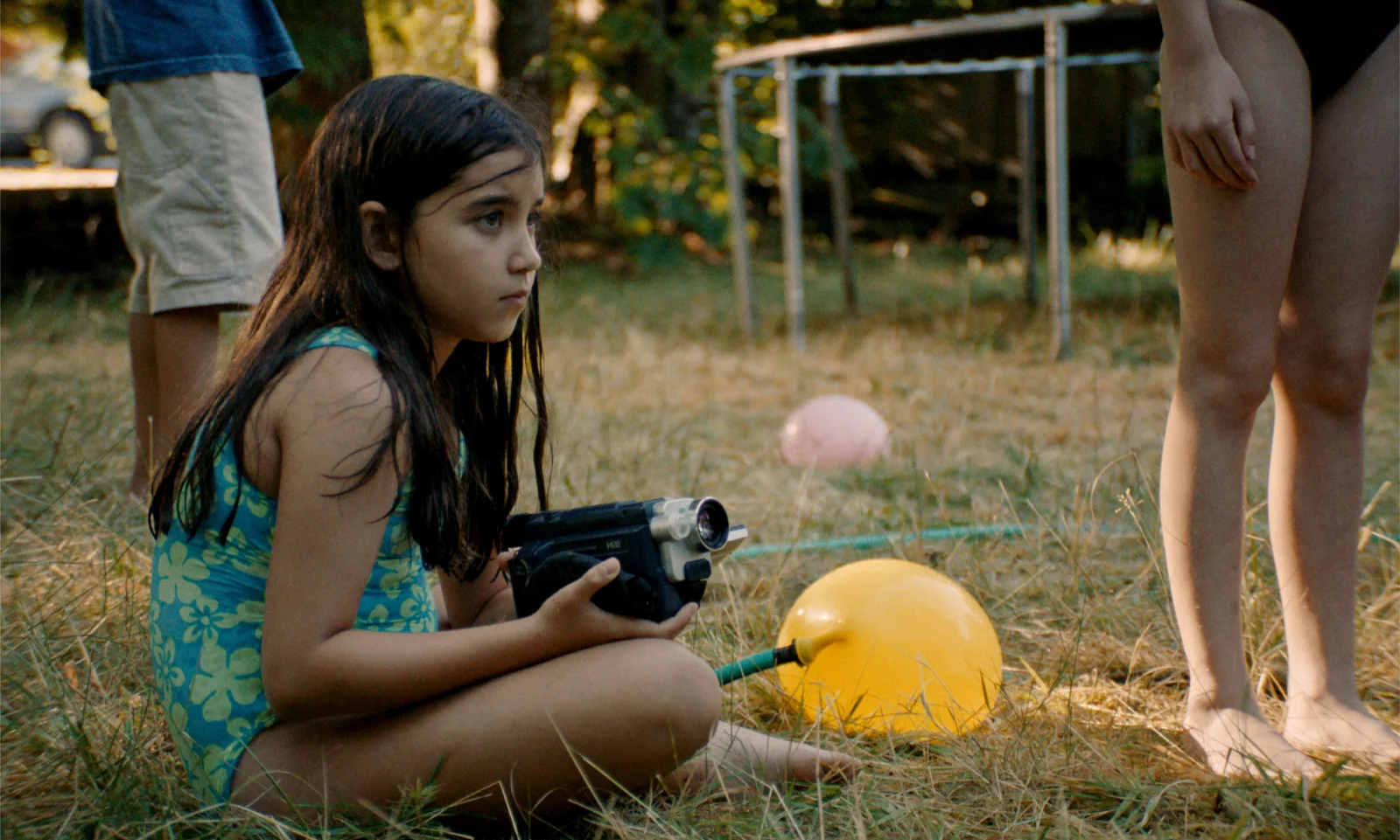 Fotograma de «Blue Heron»: una niña en bañador, sentada en la hierba con una cámara de vídeo, junto a un globo amarillo.