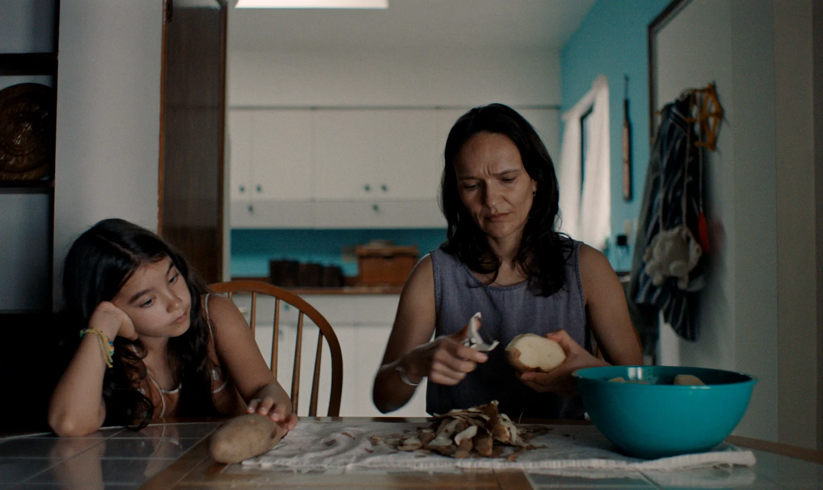 Fotograma de «Blue Heron»: una niña apoyada en la mesa de la cocina mira cómo una mujer pela patatas.