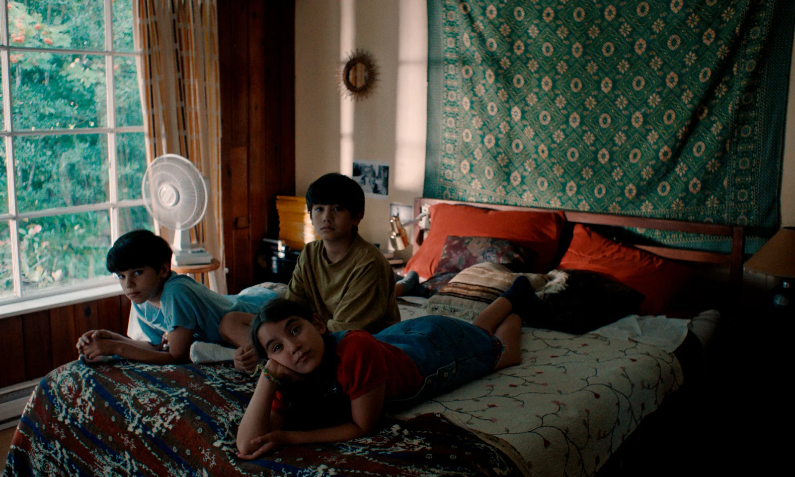 Fotograma de «Blue Heron»: tres niños tumbados en una cama miran hacia la cámara en una habitación con un gran ventanal.
