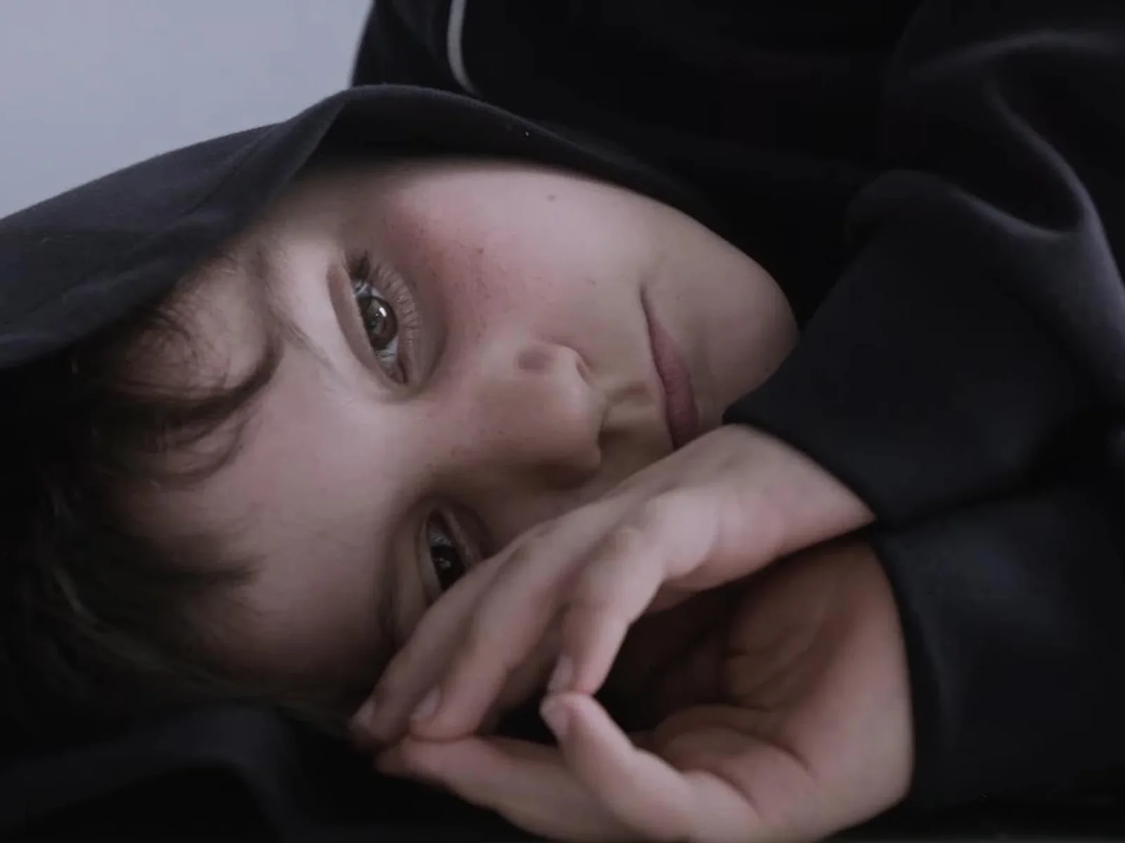 Fotograma de «On vous croit»: primer plano de un niño tumbado de lado, con capucha negra y las manos junto a la cara, mirando a cámara.