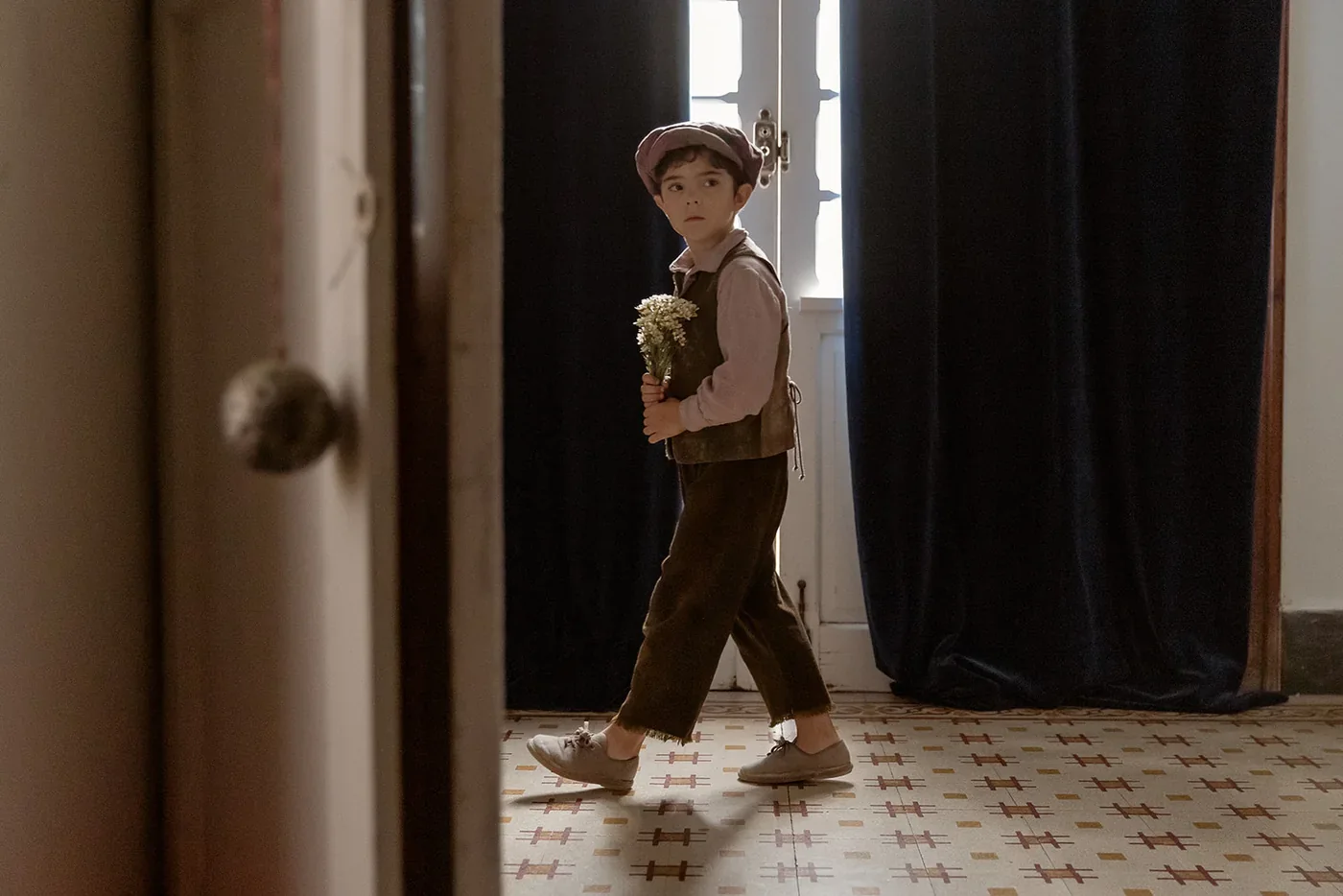 Fotograma de «Arriba tutto»: un niño con gorra y chaleco cruza una habitación con un ramo de flores en la mano.