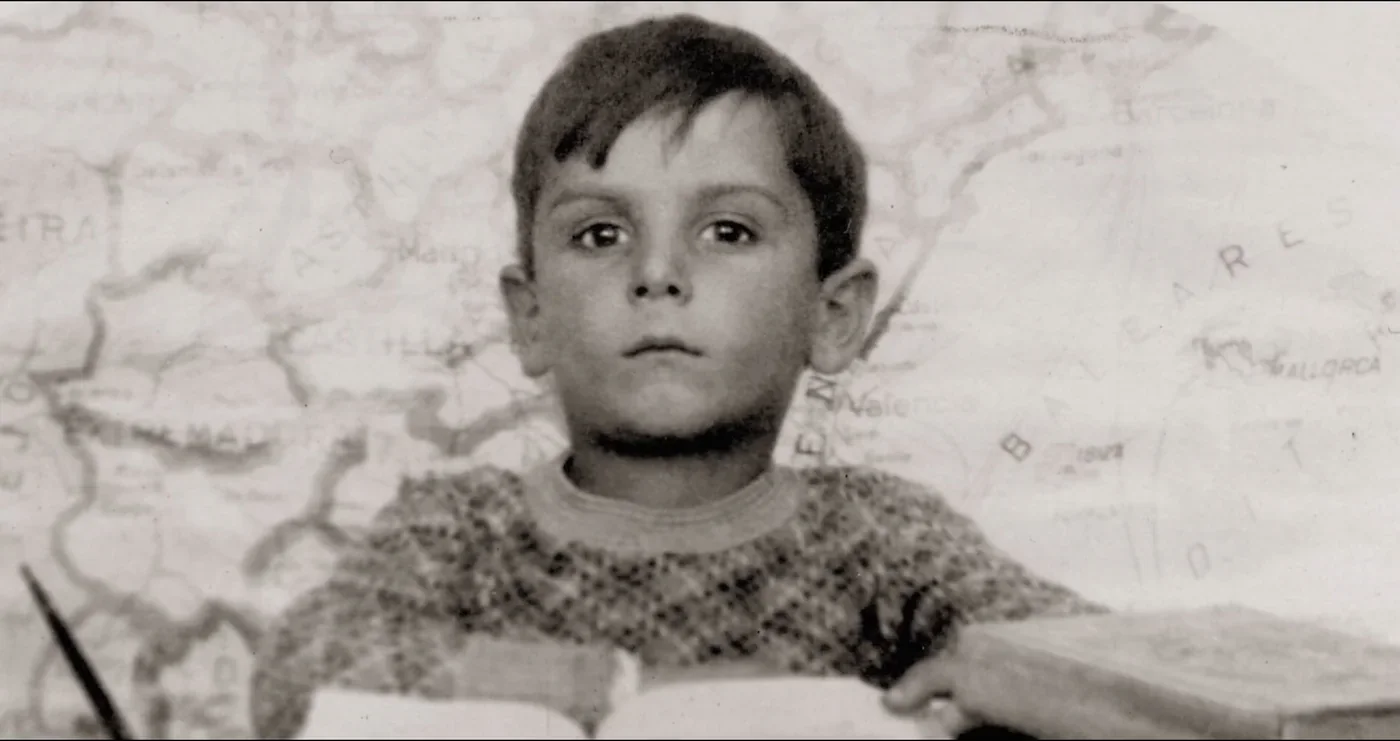Fotograma de «Carlos Saura. Ese niño de la fotografía»: fotografía en blanco y negro de un niño ante un mapa, con un libro abierto.