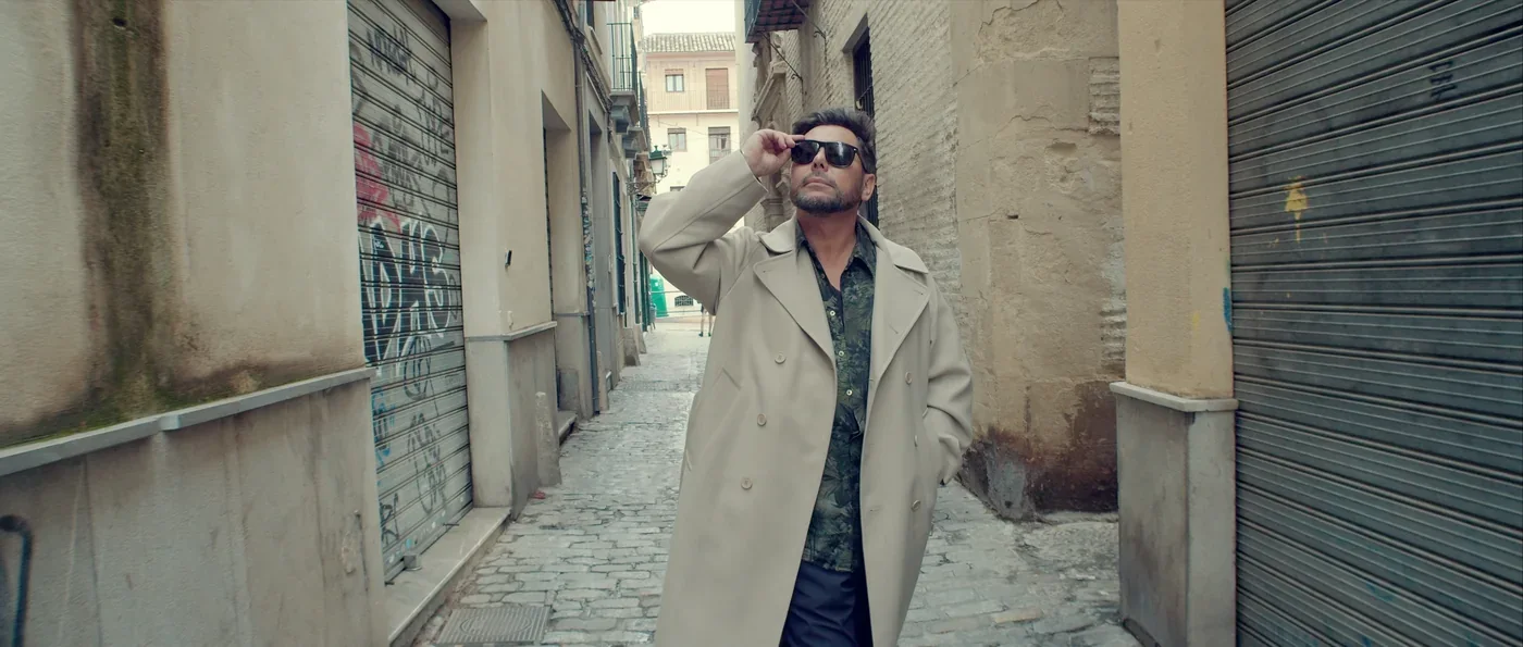 Fotograma de «Enlorquecido: solo el misterio nos hace vivir»: un hombre con gabardina y gafas de sol camina por una calle estrecha.