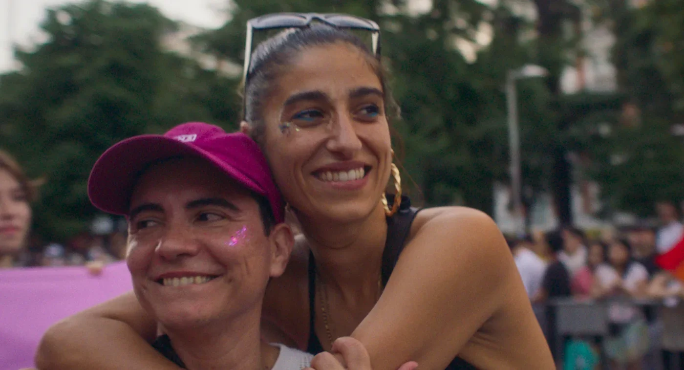 Fotograma de «Este cuerpo mío»: dos amigos se abrazan sonrientes en una manifestación; uno lleva una gorra rosa.