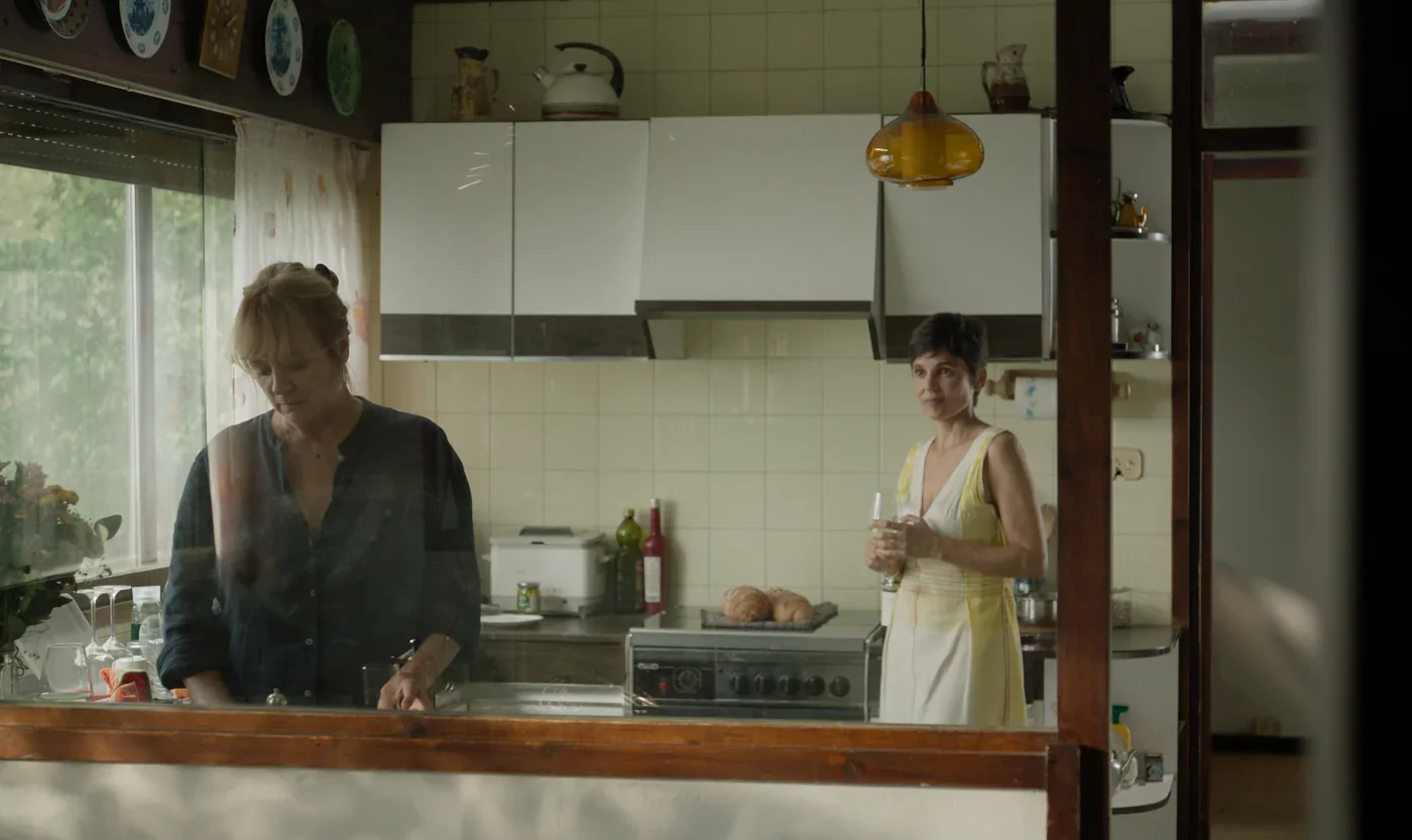 Fotograma de «Hermanas»: dos mujeres en una cocina: una prepara la comida y la otra la mira con una copa en la mano.