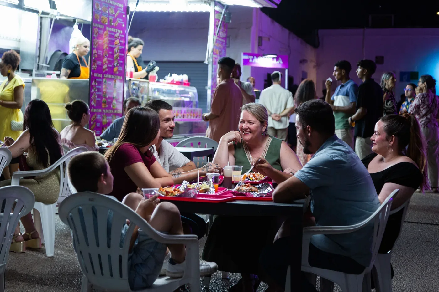 Fotograma de «Iván & Hadoum»: un grupo de personas cena y ríe en una terraza junto a un puesto de comida, de noche.
