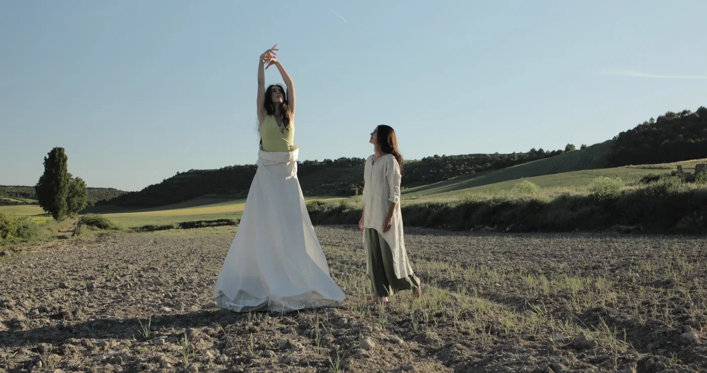 Fotograma de «Mudras. Bailando hilos invisibles»: dos mujeres en un campo: una baila con un vestido blanco y la otra la observa.
