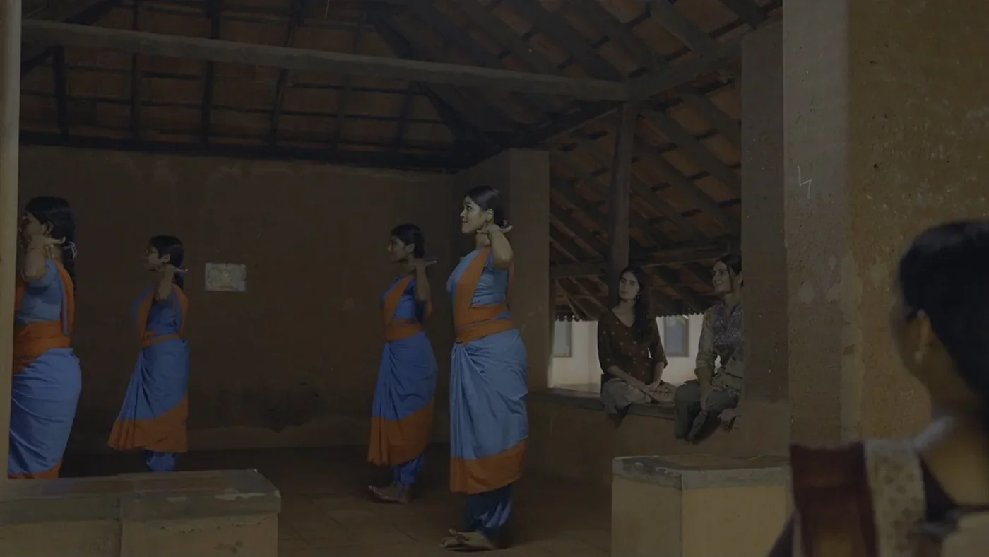 Fotograma de «Mudras. Bailando hilos invisibles»: unas bailarinas con saris azules ensayan bajo un tejado de madera.