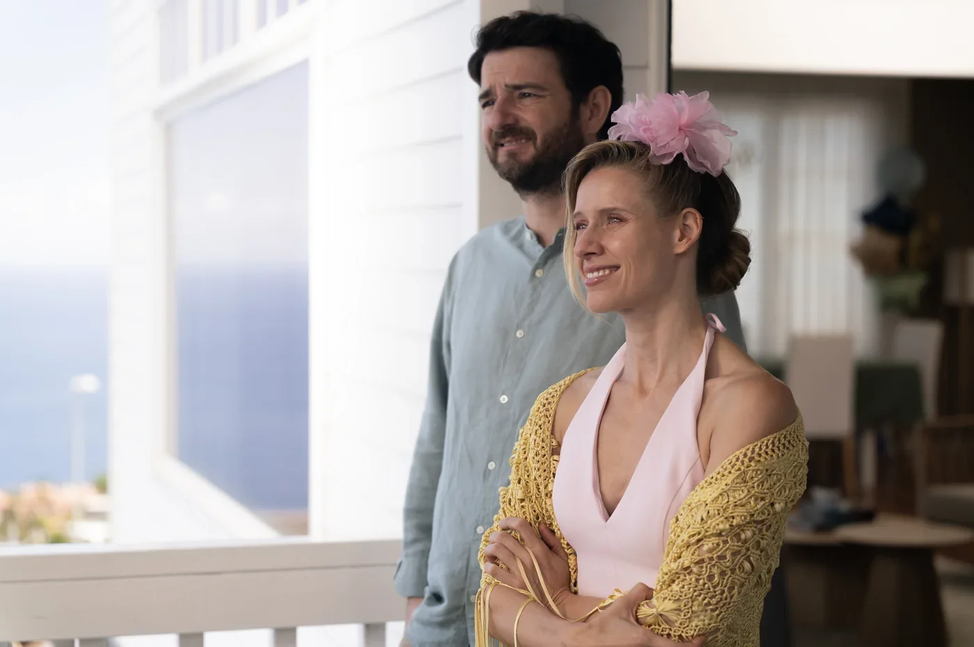Fotograma de «Ni contigo ni sin mí»: una mujer sonriente con un tocado de flores rosas y un hombre, asomados a una terraza.