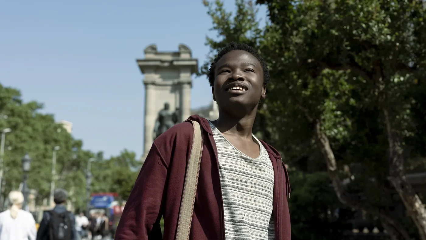 Fotograma de «Viaje al país de los blancos»: un joven sonríe mirando al cielo en una avenida arbolada con un monumento al fondo.